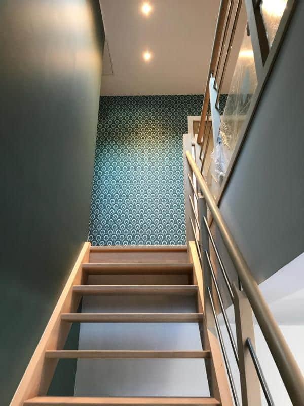 Escalier moderne avec des marches en bois clair menant à un mur accentué de papier peint à motifs géométriques bleu et blanc, éclairé par des spots au plafond.